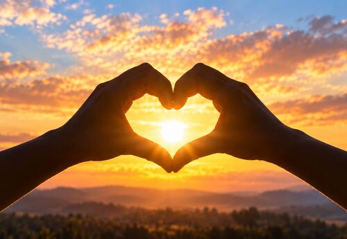 Silhouetted hands form a heart around the glowing sun against a warm, colorful sunset sky.