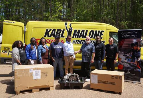 Group photo of eight adults standing outdoors in front of a bright yellow service van branded “Karl Malone Toyota.” Two men in the center are shaking hands, suggesting a partnership or donation. In front of the group are large boxed items and an exposed a