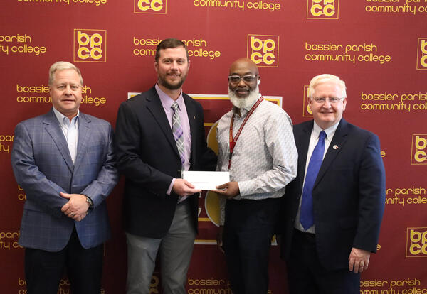 Four men standing in front of a backdrop with the BPCC logo for a check presentation