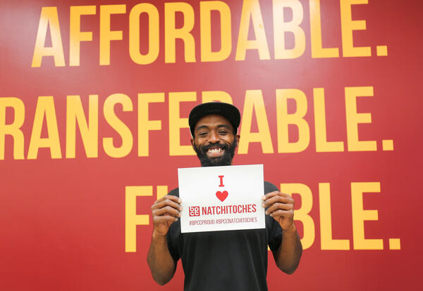 Student holding a sign that says I love BPCC Nathitoches and standing in front of a wall that says Affordable, Transferable, Flexible