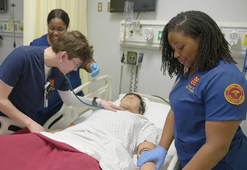 Three BPCC nursing students practice patient assessment and care using a simulation mannequin in a nursing lab.