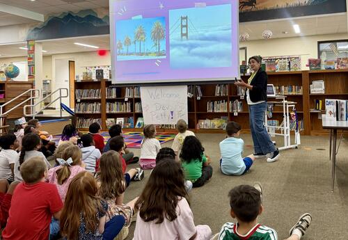 Students sitting on floor watching adult speaking at front of room