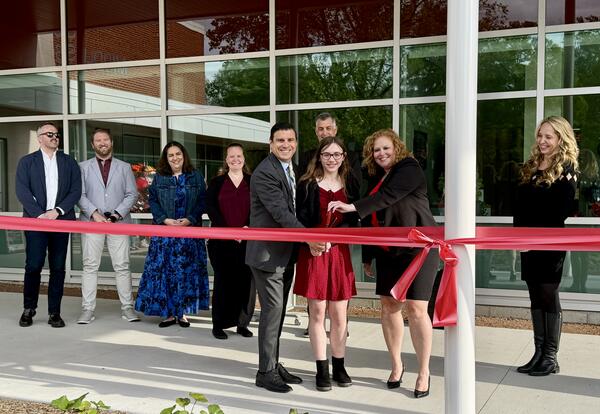 Three people dressed in business attire hold oversized scissors about to cut red ribbon with several people behind watching