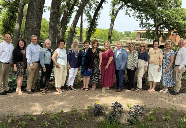 16 people standing in line on gravel path under large trees