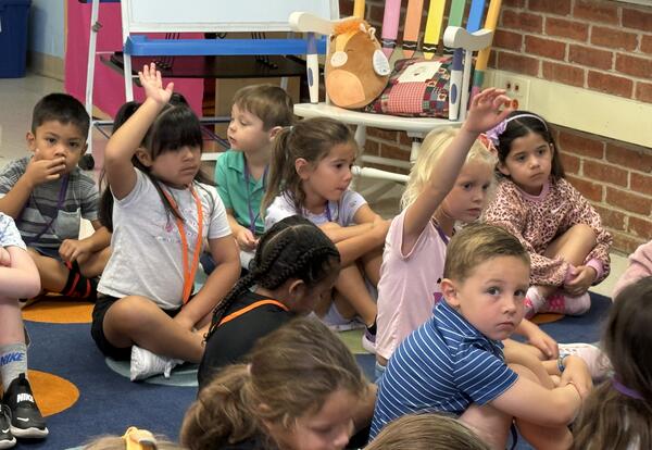 Kindergarten students sitting on classroom rug with hands raised