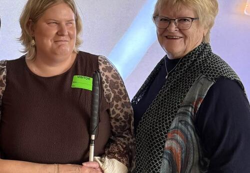 Two women stand side by side smiling. One wears a visitor badge and holds a white cane, while the other stands close beside her. Both appear warmly engaged, suggesting a classroom or school visit setting