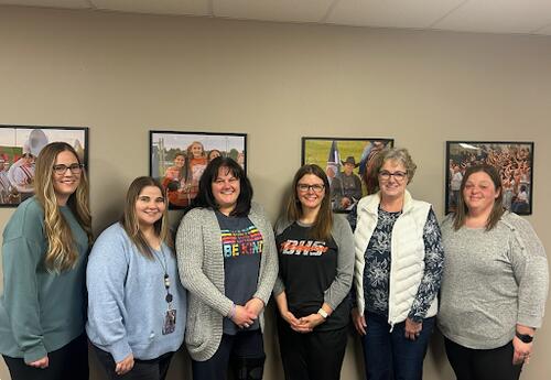 Six district social workers stand side by side in an office, smiling at the camera, with framed photos on the wall behind them