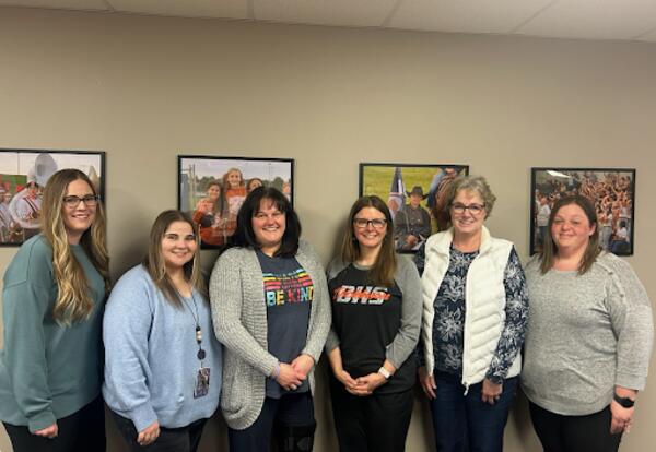 Six district social workers stand side by side in an office, smiling at the camera, with framed photos on the wall behind them