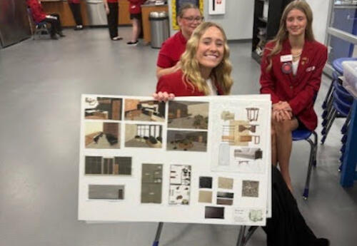 Three FCCLA students in red jackets sit in a classroom, smiling and presenting a board with interior design samples and project images.