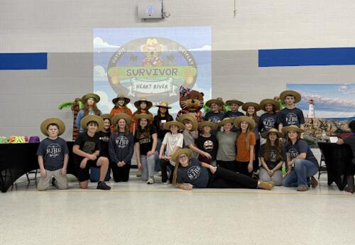 Students and family members pose together in a school gym wearing hats for a ‘Survivor Night’ event, with a themed ‘Survivor: Heart River’ logo projected behind them.