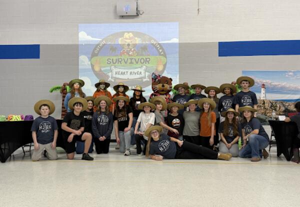 Students and family members pose together in a school gym wearing hats for a ‘Survivor Night’ event, with a themed ‘Survivor: Heart River’ logo projected behind them.