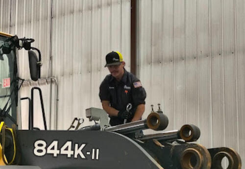 Connor in a workshop standing beside a large piece of heavy machinery labeled ‘844K-II.’ He is wearing a dark uniform, gloves, and a black cap, and appears to be inspecting or adjusting a metal component on the equipment. The background shows a tall indus