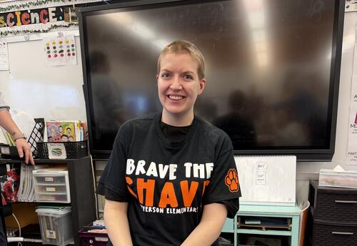 Miss Ramsey smiling with a freshly shaved head sits on a chair in a classroom, wearing a ‘Brave the Shave’ T-shirt. Hair clippings are visible on a blue tarp beneath them, with classroom materials and a large screen in the background
