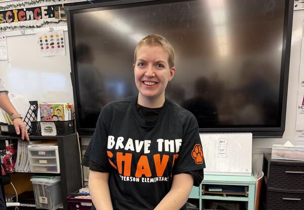 Miss Ramsey smiling with a freshly shaved head sits on a chair in a classroom, wearing a ‘Brave the Shave’ T-shirt. Hair clippings are visible on a blue tarp beneath them, with classroom materials and a large screen in the background