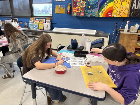  Gemini said Three teenage girls sit at a table in a brightly colored art classroom, focused on their drawings. They are using charcoal and colored pencils to create realistic 3D sphere illustrations on blue and yellow paper. The background features blue walls adorned with student artwork, including a "Starry Night" recreation and colorful murals.