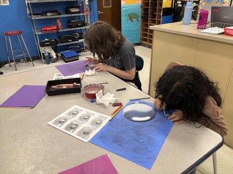 Two female students lean over a gray classroom table, carefully working on art projects. They are using white and black charcoal on blue and purple paper to draw spheres with realistic highlights and shadows. Instructional worksheets, pencils, and blending tissues are scattered across the table. The background shows a classroom setting with blue walls, storage shelves, and student-painted lockers.