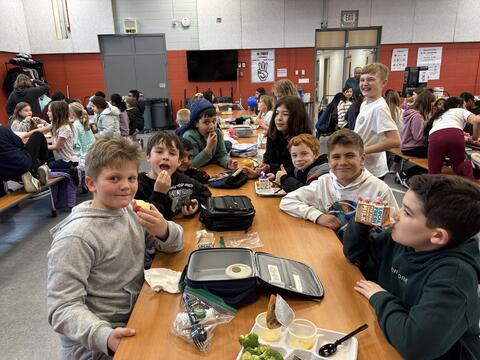 A group of elementary-aged boys sit together at a long wooden table in a busy school cafeteria. They are smiling and looking at the camera while eating their lunches, which include apples, broccoli, and chocolate milk cartons. The background is filled with other students eating and talking in a room with bright red walls.