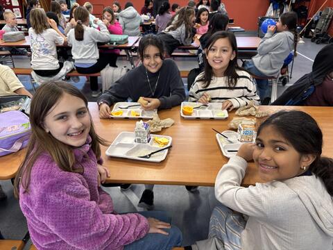 Four young elementary-aged girls smile at the camera while sitting together at a wooden cafeteria table. They are eating school lunches consisting of fruit, rice, and milk cartons on white divided trays. The background is a busy school lunchroom filled with other students at long tables.