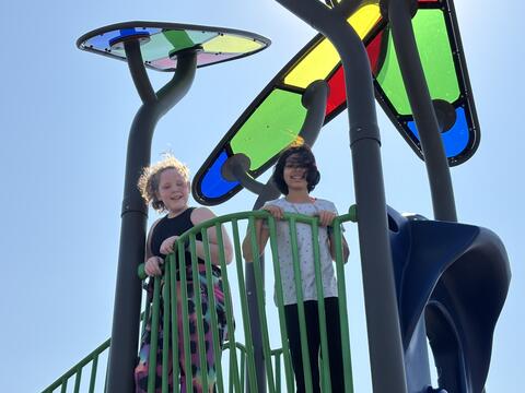 A low-angle photo of two young girls smiling on a colorful playground. They stand on a green bridge, looking down at the camera. The girl on the left wears a black top and tie-dye leggings; the girl on the right wears a patterned white shirt. Modern sun shades with blue, green, and red panels tower above them against a clear, blue sky.