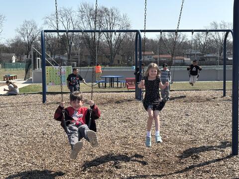Two young children smile while swinging on a black belt swing set at a sunny outdoor playground. The boy on the left wears a red shirt and a red and black plaid jacket, while the girl on the right wears a black floral dress. Other children play in the background near a blue picnic table and a communication board, with bare trees and houses visible under a clear sky.
