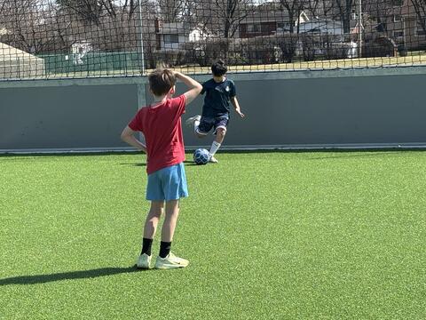 Two young boys play soccer on a bright green artificial turf field on a sunny day. One boy in a dark blue shirt is mid-kick with a blue patterned soccer ball, while another boy in a red t-shirt and blue shorts watches from the foreground with his hand on his head. A grey wall and a black safety net border the field, with houses and bare trees visible in the distance.