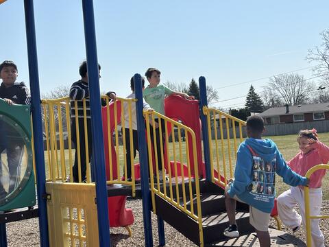 A group of elementary school children play on a colorful primary-colored playground structure under a clear blue sky. Several boys stand on the elevated platforms behind yellow railings, while a boy in a blue hoodie and a girl in a pink sweatshirt climb the stairs in the foreground. The playground sits on a woodchip surface with a grassy field and houses in the background.