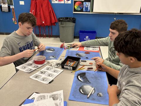 Three teenage boys sit at a classroom table working on art projects. They are using pencils and charcoal to draw realistic, shaded water droplets on bright blue paper, following instructional worksheets. Red art smocks hang on the blue wall in the background of the art studio.