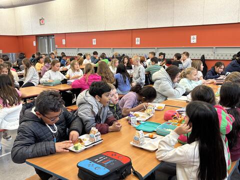  Gemini said A large group of elementary school students sit at long wooden tables in a lively school cafeteria. The children are seen eating lunch from white divided trays and personal lunchboxes, chatting with one another. The room features high ceilings with acoustic panels and a prominent red accent wall lined with coat hooks.