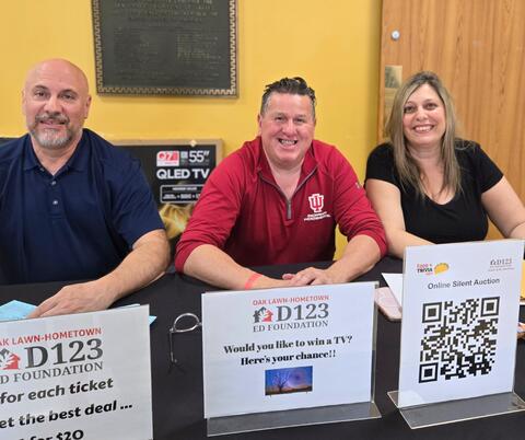 Three people sit behind a table promoting a D123 Foundation TV raffle with signs and a QR code for an online silent auction.