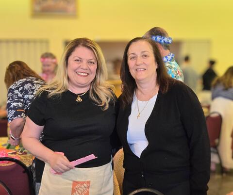 Two women smiling and posing together indoors at a social gathering with other people in the background.