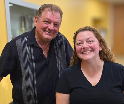 Two people standing indoors against a yellow wall, one wearing a black shirt with vertical stripes and the other in a plain black V-neck shirt. 