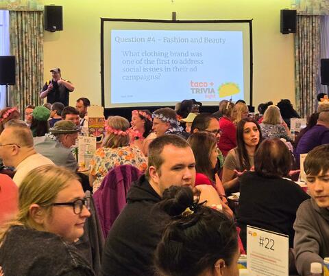 Crowded room with people participating in a taco trivia night, a host stands near a screen showing a question.
