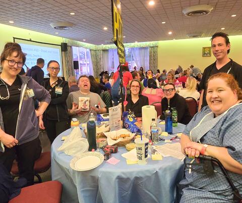 Group of people dressed as characters from the show The Pitt gathered around a table with snacks and drinks at a community event in a large room.