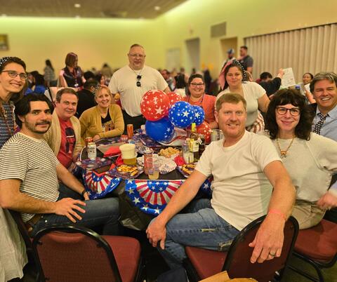 Group of people gathered around a table with snacks and drinks in a community hall setting. 