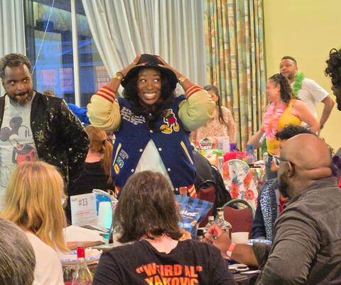 A lively indoor gathering with people seated and standing around tables, engaging in conversation and activities.