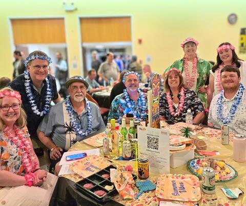 Group of people wearing Hawaiian leis and tropical shirts seated around a table with drinks and themed party decorations indoors.