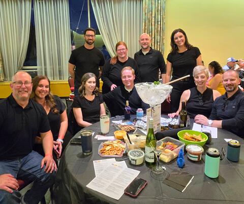 Group of friends gathered around a table with snacks, drinks, and a large ice-filled cocktail glass in an indoor setting at night.