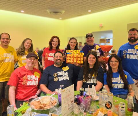 Group of people wearing colorful 'Come On Down' shirts posing around a table filled with snacks and drinks at an indoor event.