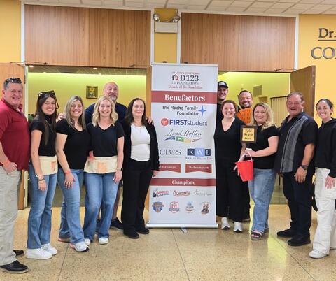 Group of event volunteers standing indoors around a banner listing sponsors and benefactors for a community Taco & Trivia event