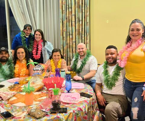Group of people wearing Hawaiian leis and tropical shirts seated around a table with drinks and themed party decorations indoors.