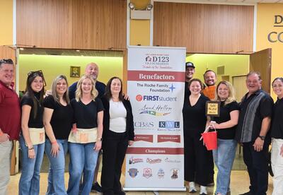 Group of event volunteers standing indoors around a banner listing sponsors and benefactors for a community Taco & Trivia.