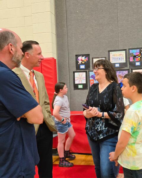 A group of adults and children engaged in conversation inside a school gym with student artwork displayed on the wall behind them.