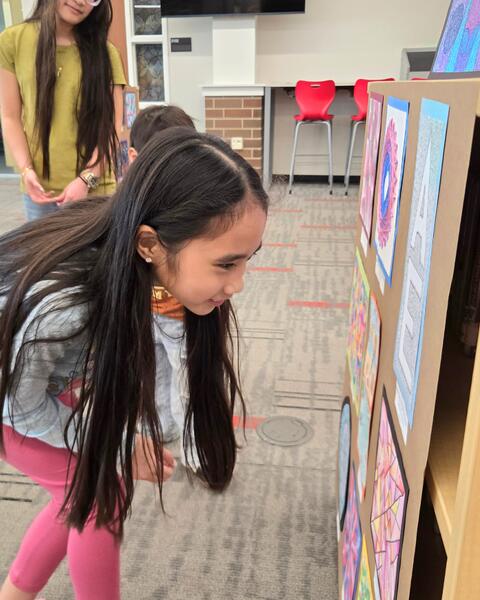 Young girl closely examining colorful artwork displayed on a board in a classroom setting.