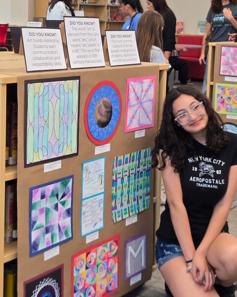 Student sitting beside a display board featuring colorful geometric and abstract art pieces in a classroom setting.