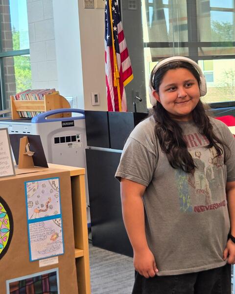 Student with headphones smiling beside a display of artwork in the library