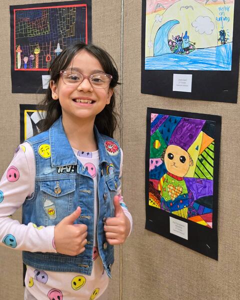 Child wearing a denim vest with colorful patches giving thumbs up in front of vibrant student artwork displayed on a beige wall.
