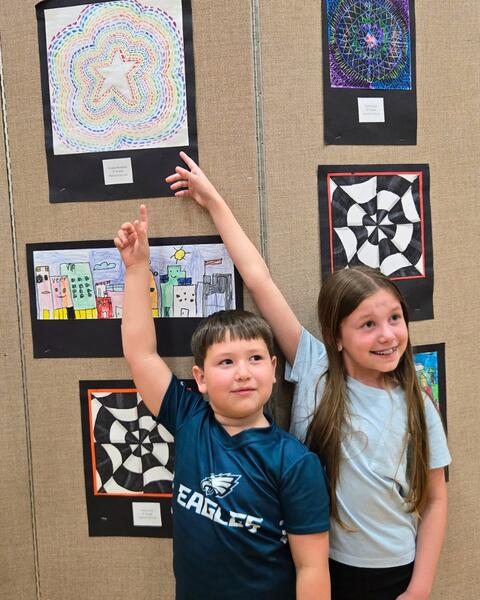 Two children pointing excitedly at colorful artwork displayed on a beige fabric-covered wall in a school or gallery setting.