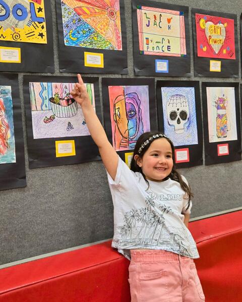 Child pointing excitedly at colorful student artwork displayed on a gray wall in a school setting.