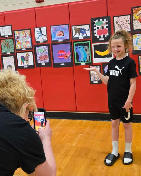 Child in black Puma outfit presenting colorful artwork displayed on a red gym wall while an adult photographs them with a smartphone.