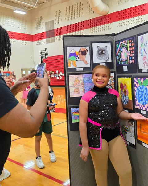 Young girl in a sparkly black and pink dance costume posing next to colorful artwork displayed on a panel in a school gymnasium.